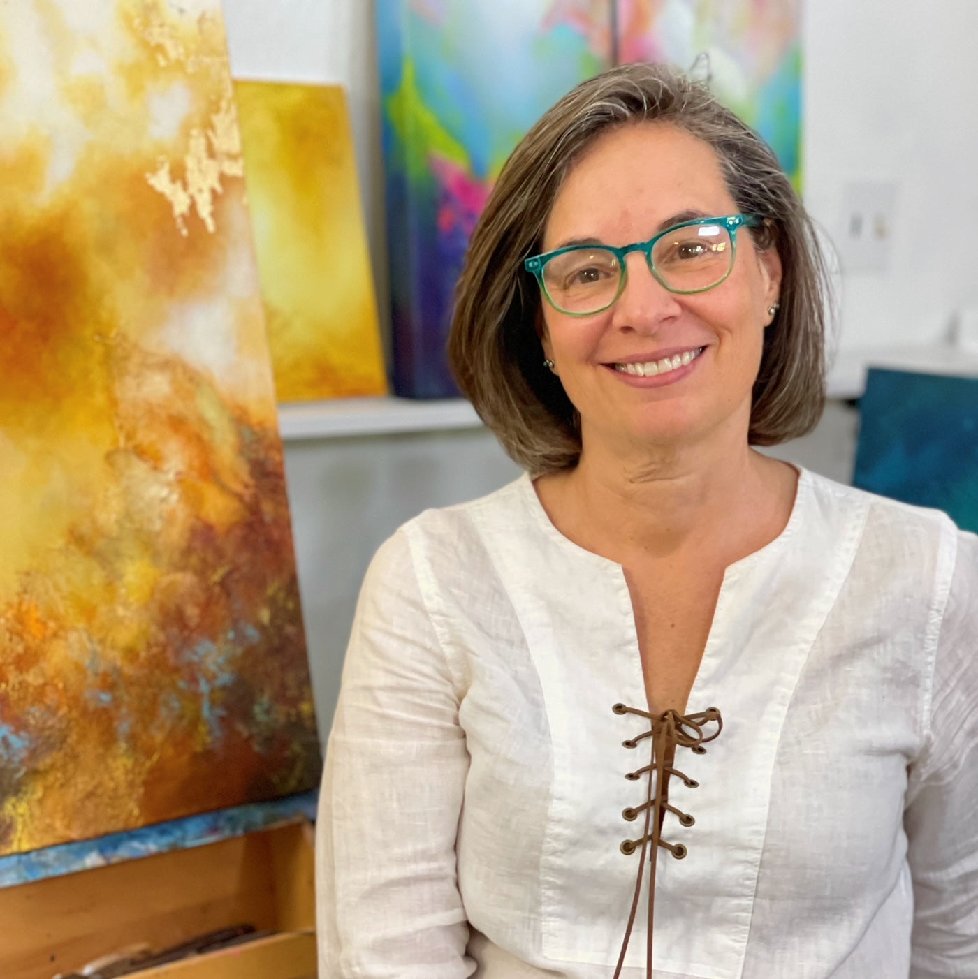 Jennifer Tepper sits in her New Mexico studio, wearing aqua blue glasses and wearing a white blouse with leather lacing up the front, in front of several of her abstract paintings. Jennifer Tepper Fine Art