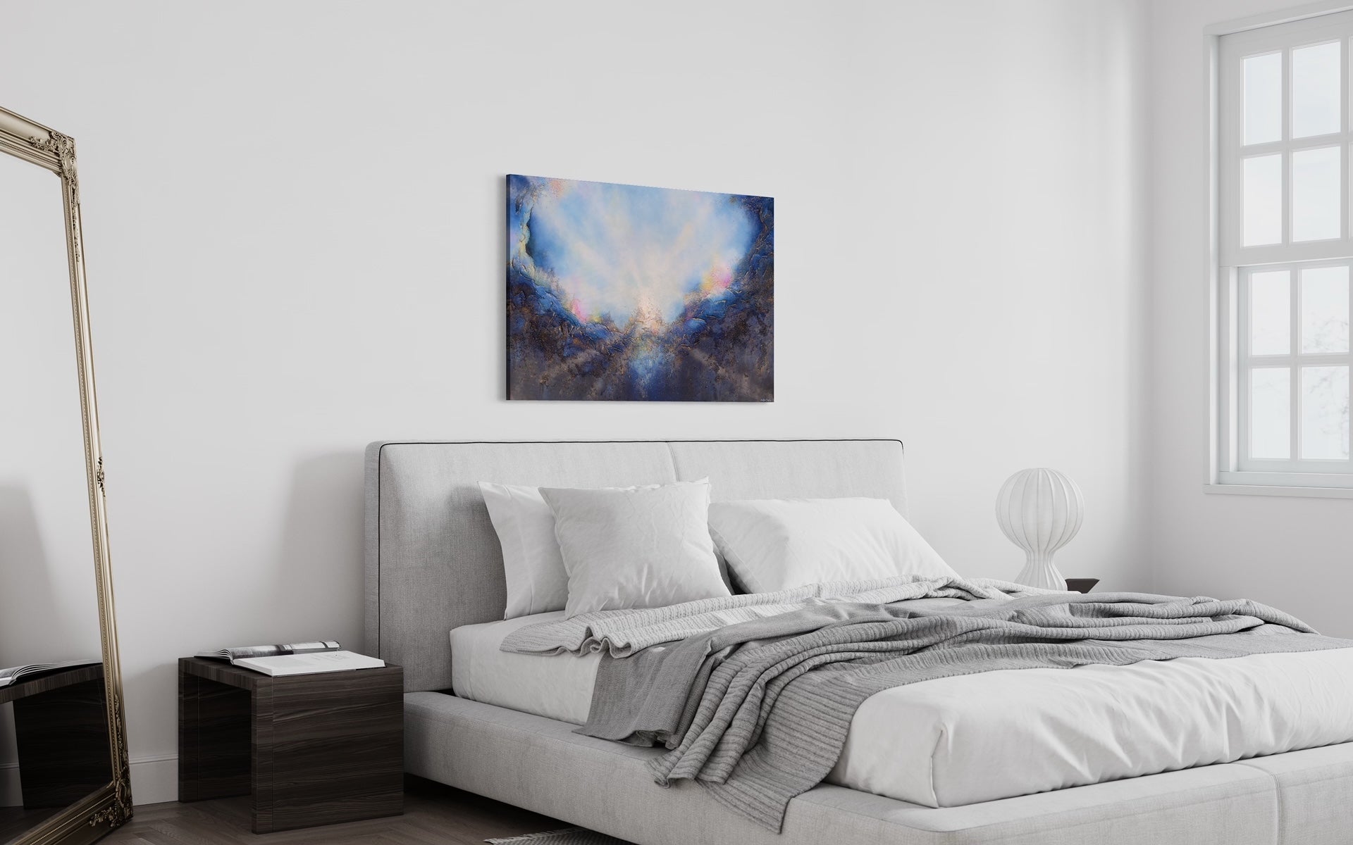Soul's Return, hanging in a bedroom, over a bed with gray padded headboard and a gray comforter and crisp white sheets, white walls and deep brown bedside table.  A textured painting with light rays shining out from the center representing the soul's return to one's self, or the universe. Blue, pink, white, dark blue, textured painting. Ephemeral art, art with meaning, soul artist. Jennifer Tepper Fine Art.
