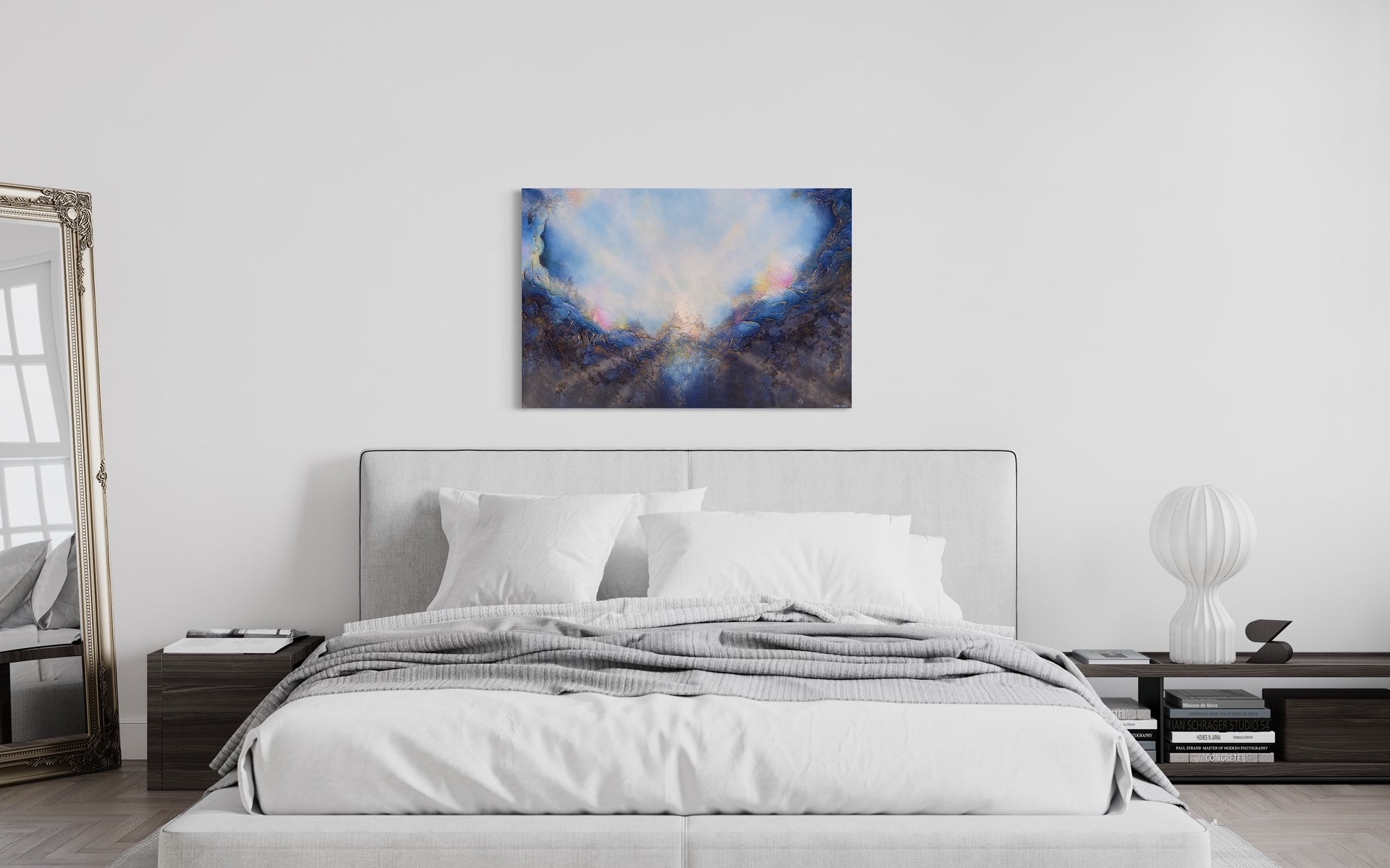Soul's Return, hanging in a bedroom, over a bed with gray padded headboard and a gray comforter and crisp white sheets, white walls and deep brown bedside table.  A textured painting with light rays shining out from the center representing the soul's return to one's self, or the universe. Blue, pink, white, dark blue, textured painting. Ephemeral art, art with meaning, soul artist. Jennifer Tepper Fine Art.